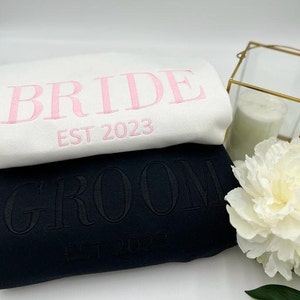 May include: Two sweatshirts, one white and one black, are stacked. The white sweatshirt has "BRIDE EST 2023" embroidered in pink. The black sweatshirt has "GROOM EST 2023" embroidered in black. The sweatshirts are likely for a wedding.