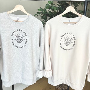 May include: Two sweatshirts with a floral design and the text "Consider how the wildflowers grow" and a bible verse reference. The sweatshirt on the left is gray and the sweatshirt on the right is cream.