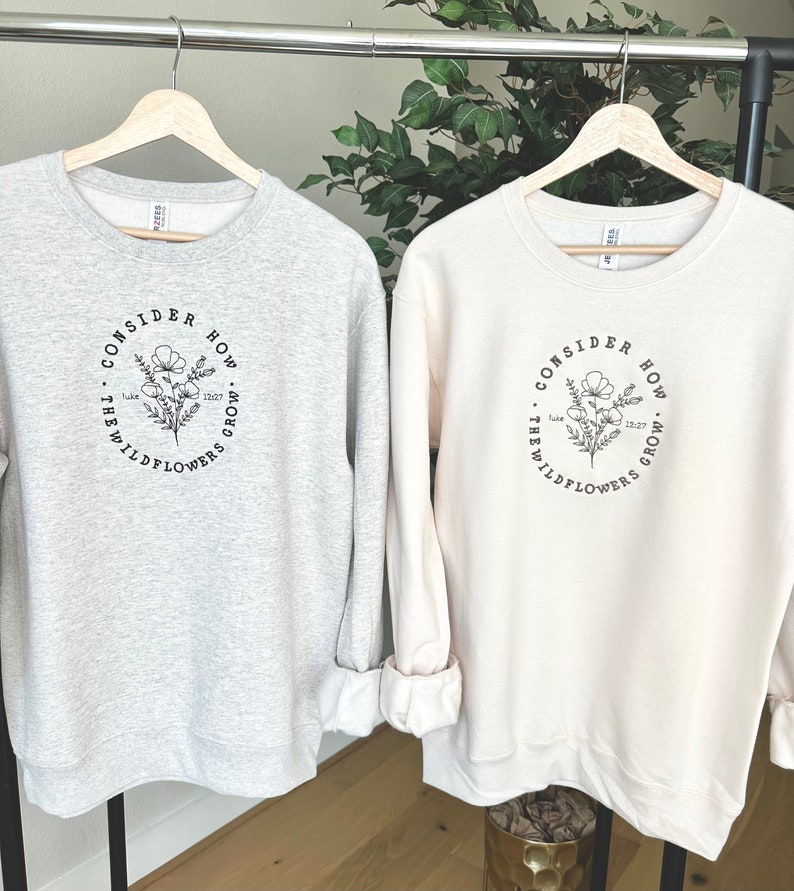 May include: Two sweatshirts with a floral design and the text "Consider how the wildflowers grow" and a bible verse reference. The sweatshirt on the left is gray and the sweatshirt on the right is cream.