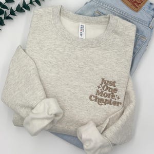 May include: A light heather gray crewneck sweatshirt featuring brown embroidered text on the front right chest that reads Just One More Chapter with small star accents. The sweatshirt has long sleeves with ribbed cuffs and a Jerzees brand label.