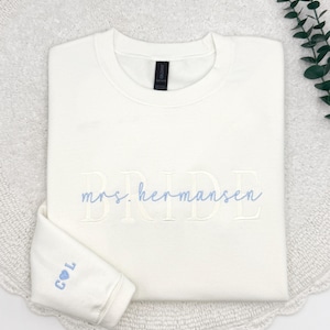 May include: Ivory-colored sweatshirt with the word "BRIDE" embroidered in a light color. The name "mrs. hermansen" is embroidered in a light blue script. The sleeve cuff displays the initials "C & L" with a heart symbol.