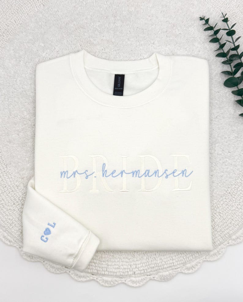 May include: Ivory-colored sweatshirt with the word "BRIDE" embroidered in a light color. The name "mrs. hermansen" is embroidered in a light blue script. The sleeve cuff displays the initials "C & L" with a heart symbol.