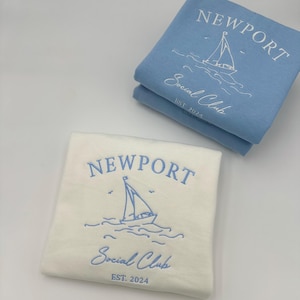 May include: Two sweatshirts with the text "Newport Social Club Est. 2024" and a sailboat graphic. One sweatshirt is white with blue embroidery and the other is light blue with white embroidery.