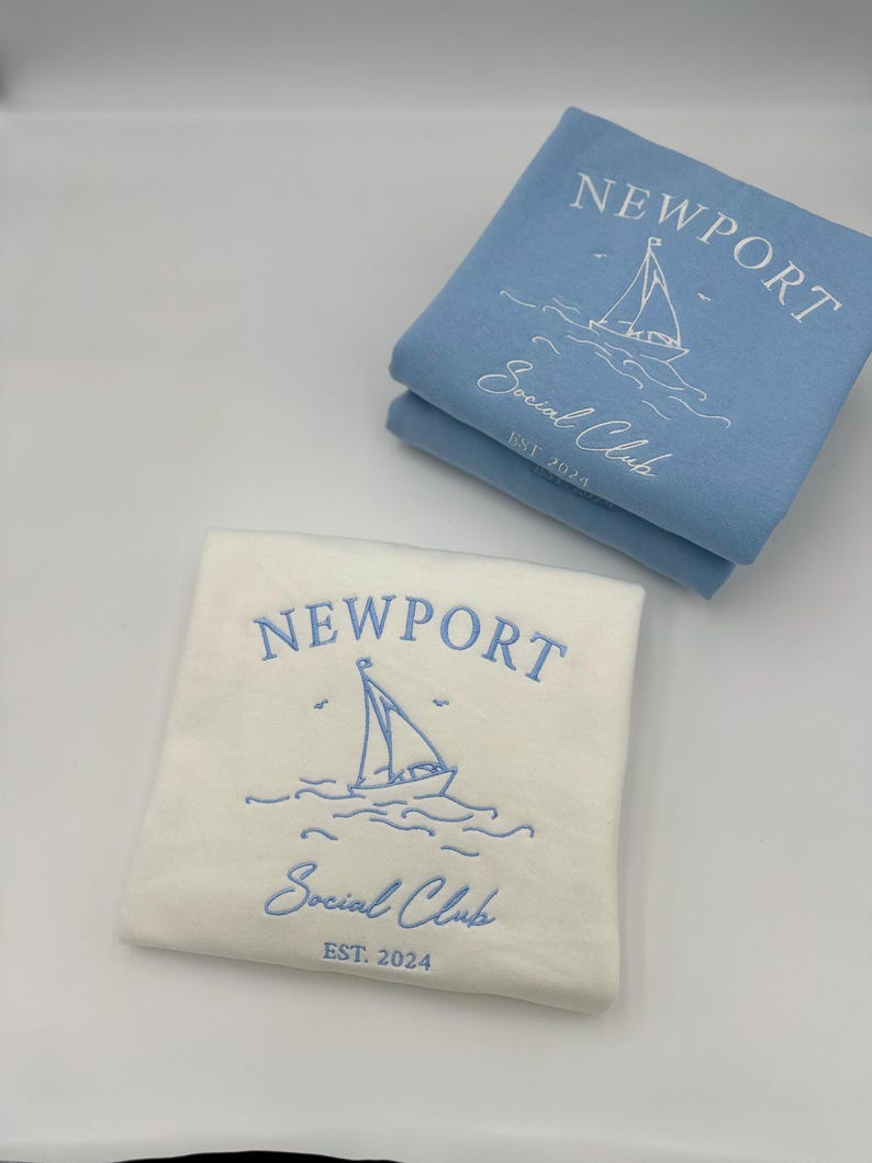 May include: Two sweatshirts with the text "Newport Social Club Est. 2024" and a sailboat graphic. One sweatshirt is white with blue embroidery and the other is light blue with white embroidery.