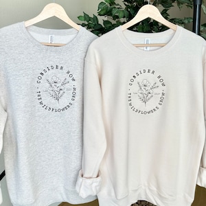 May include: Two sweatshirts with a floral design and the text "Consider how the wildflowers grow."  The text is in a circle around the floral design. One sweatshirt is light gray and the other is cream.