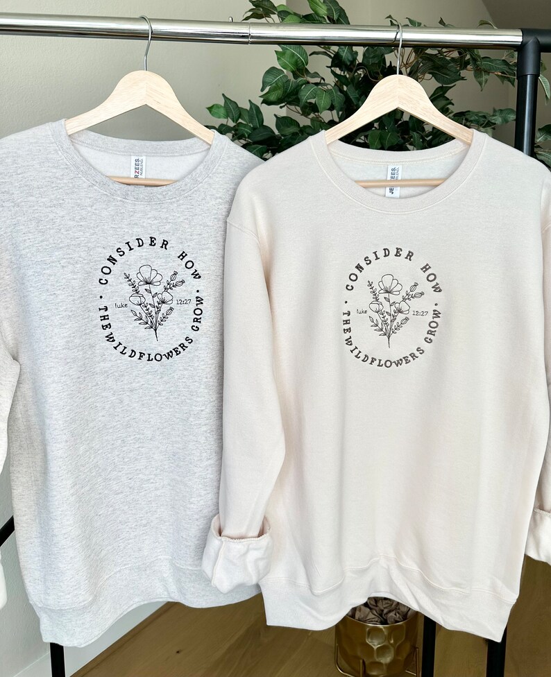 May include: Two sweatshirts with a floral design and the text "Consider how the wildflowers grow."  The text is in a circle around the floral design. One sweatshirt is light gray and the other is cream.
