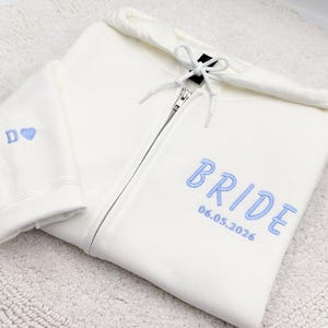 May include: Cream-colored zip-up hoodie with light blue embroidered text that reads "BRIDE" and "06.05.2026". The left sleeve has the letter "D" and a heart. The hoodie has a drawstring hood and a silver zipper.