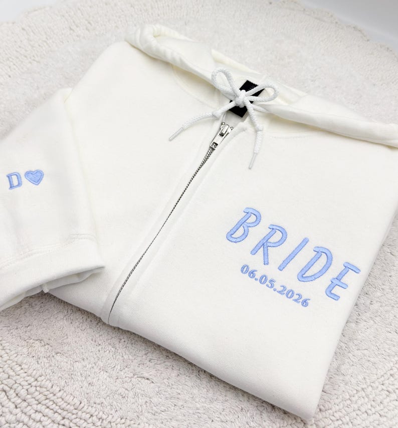 May include: Cream-colored zip-up hoodie with light blue embroidered text that reads "BRIDE" and "06.05.2026". The left sleeve has the letter "D" and a heart. The hoodie has a drawstring hood and a silver zipper.