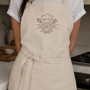May include: A beige apron featuring an embroidered design of a chef hat, a whisk, and a spatula crossed over each other. Below the utensils, the text CUSTOM TEXT is centered within a horizontal line, with a small leaf motif at the very bottom.