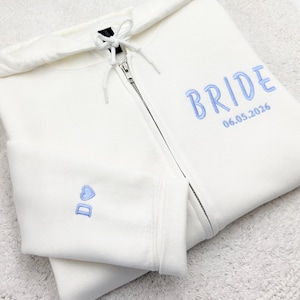 May include: A white zip-up hoodie with a drawstring hood. The word "BRIDE" is embroidered in light blue on the front, along with the date "06.05.2026". The left sleeve features a small heart and the initials "D O" in light blue.