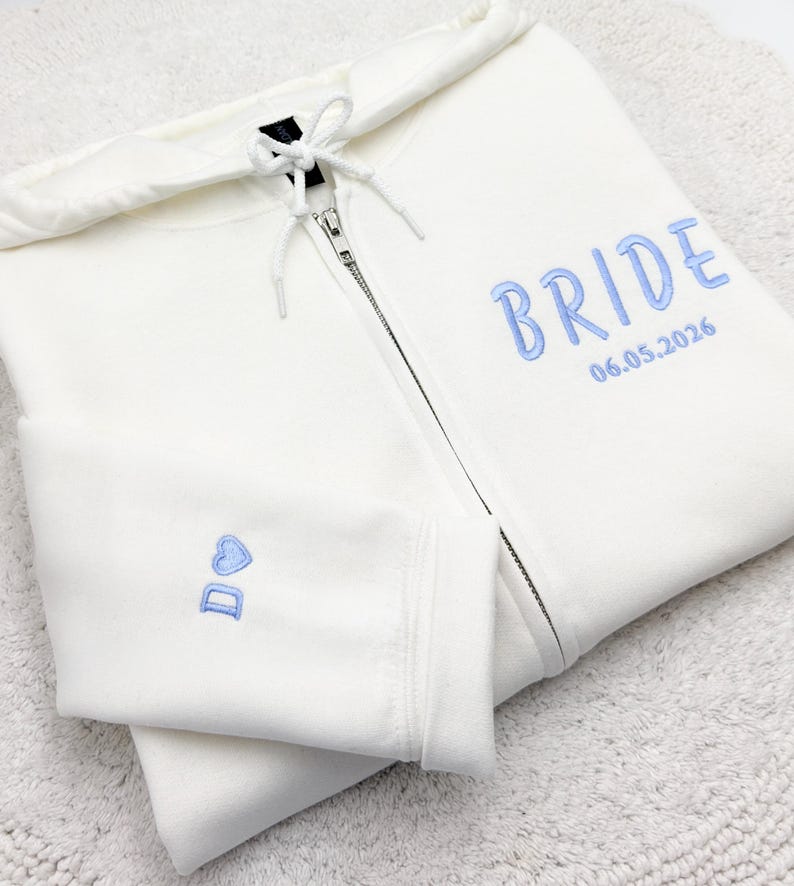 May include: A white zip-up hoodie with a drawstring hood. The word "BRIDE" is embroidered in light blue on the front, along with the date "06.05.2026". The left sleeve features a small heart and the initials "D O" in light blue.