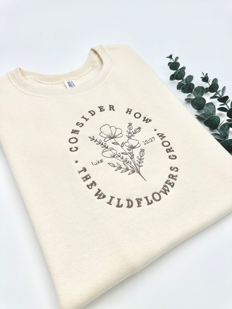 May include: A cream-colored sweatshirt with a brown embroidered design that says "Consider how the wildflowers grow" with a floral illustration and the text "Luke 12:27".