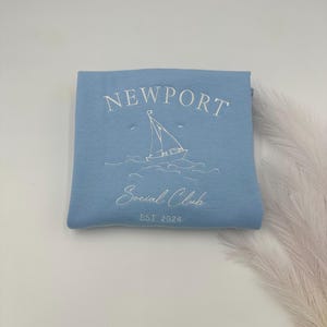 May include: A light blue sweatshirt with the text "NEWPORT Social Club EST. 2024" and a white sailboat graphic.