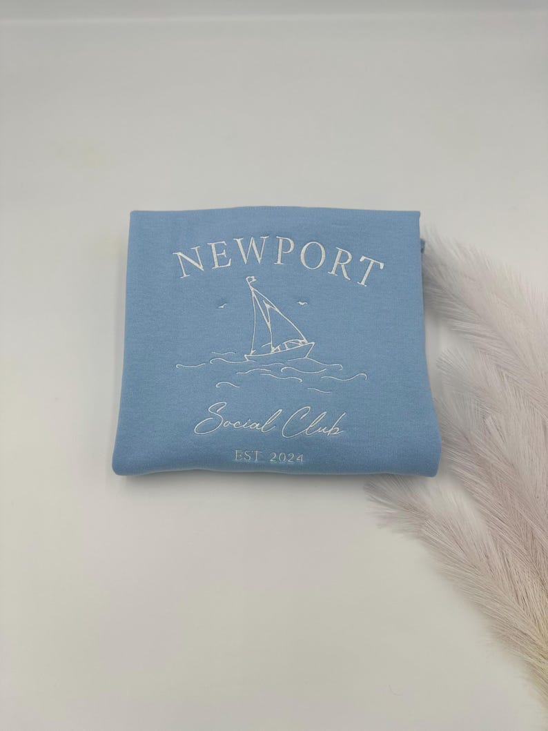 May include: A light blue sweatshirt with the text "NEWPORT Social Club EST. 2024" and a white sailboat graphic.