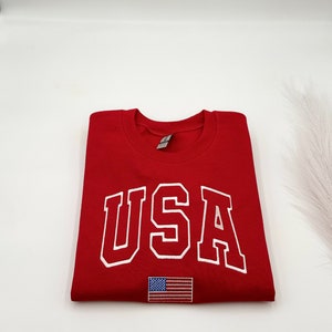 Puede incluir: Una sudadera roja doblada con la palabra "USA" en letras blancas. Debajo del texto hay una peque&ntilde;a bandera americana bordada. La sudadera est&aacute; hecha de un material suave.