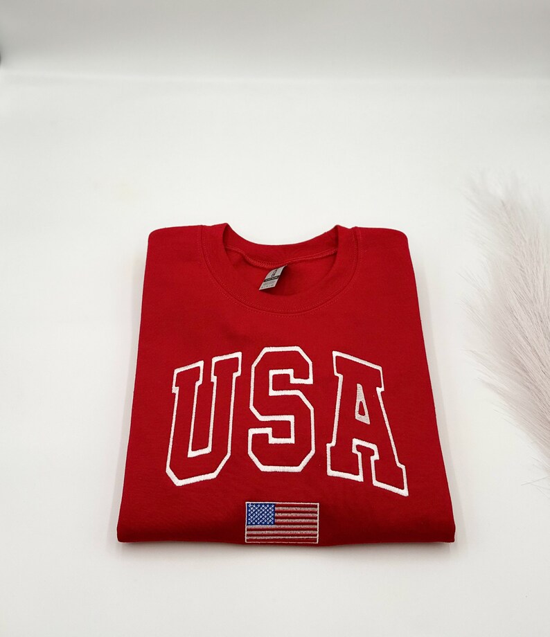 Puede incluir: Una sudadera roja doblada con la palabra "USA" en letras blancas. Debajo del texto hay una peque&ntilde;a bandera americana bordada. La sudadera est&aacute; hecha de un material suave.