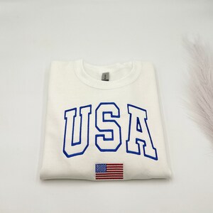 Puede incluir: Sudadera color crema con la palabra "USA" bordada en azul, arqueada sobre el pecho. Una peque&ntilde;a bandera americana est&aacute; bordada debajo del texto. La sudadera est&aacute; doblada cuidadosamente.