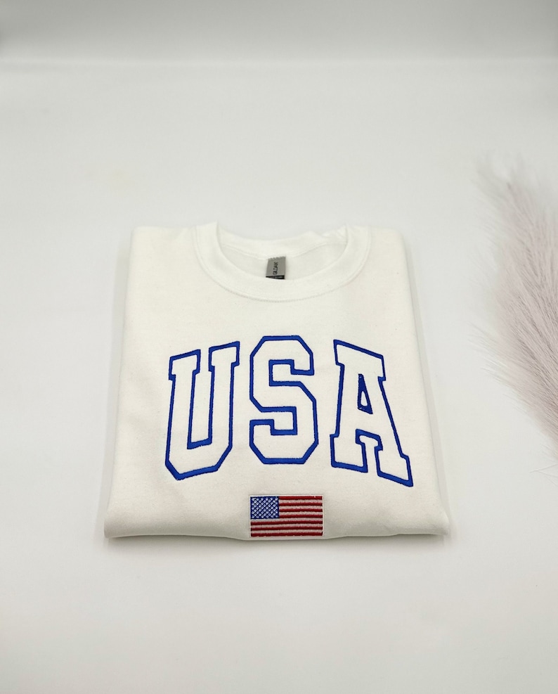 Puede incluir: Sudadera color crema con la palabra "USA" bordada en azul, arqueada sobre el pecho. Una peque&ntilde;a bandera americana est&aacute; bordada debajo del texto. La sudadera est&aacute; doblada cuidadosamente.