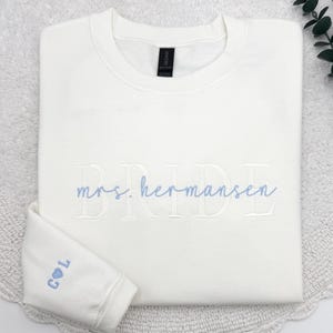 May include: A folded, off-white sweatshirt with the word "BRIDE" embroidered in light blue script. The name "mrs. hermansen" is embroidered below. The left sleeve cuff features the initials "C L" with a heart symbol, also in light blue.