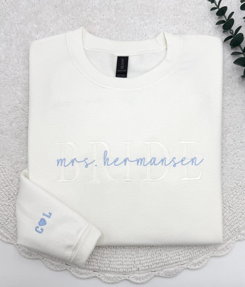 May include: A folded, off-white sweatshirt with the word "BRIDE" embroidered in light blue script. The name "mrs. hermansen" is embroidered below. The left sleeve cuff features the initials "C L" with a heart symbol, also in light blue.