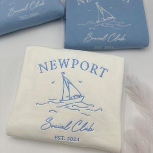 May include: Two white sweatshirts with blue embroidery. The sweatshirts feature the text "NEWPORT Social Club EST. 2024" and a sailboat graphic.