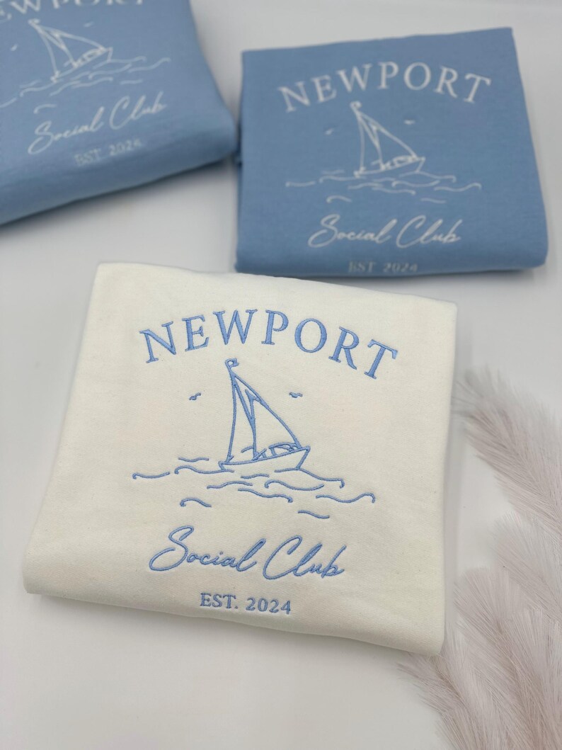 May include: Two white sweatshirts with blue embroidery. The sweatshirts feature the text "NEWPORT Social Club EST. 2024" and a sailboat graphic.