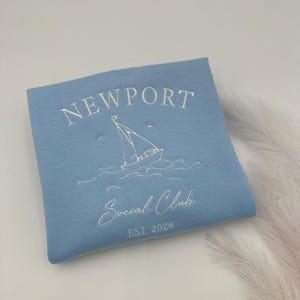 May include: Light blue sweatshirt with white embroidered text that reads "NEWPORT Social Club EST 2024" and a sailboat graphic.