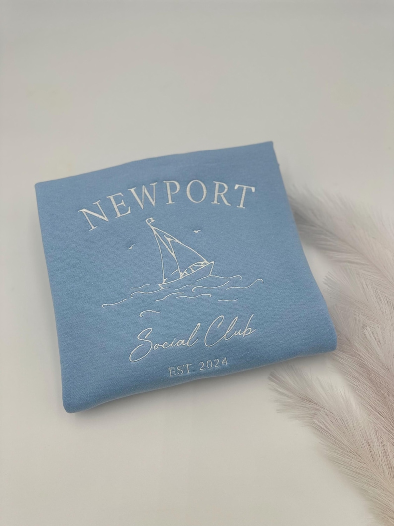 May include: Light blue sweatshirt with white embroidered text that reads "NEWPORT Social Club EST 2024" and a sailboat graphic.