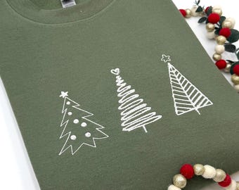 May include: An olive green crewneck sweatshirt featuring a white graphic print of three distinct stylized Christmas trees. The first tree has circular ornaments, the second has a spiral design, and the third has horizontal stripes with a star topper.