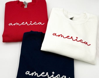 May include: Three crewneck sweatshirts in red, white, and navy blue. Each sweatshirt has the word "america" embroidered in a cursive font. The red and navy sweatshirts have white embroidery, while the white sweatshirt has red embroidery.