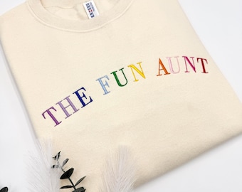 Puede incluir: Sudadera color crema con la frase bordada "THE FUN AUNT" en colores arcoíris. Esta prenda informal está hecha de un material suave. La imagen incluye vegetación decorativa.