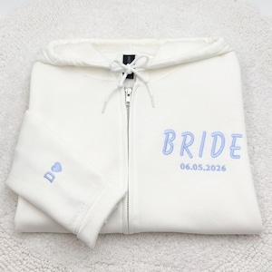 May include: A white zip-up hoodie with the word "BRIDE" embroidered in light blue on the front. The date "06.05.2026" is embroidered below the word. A small blue heart and the letter "D" are embroidered on the sleeve.