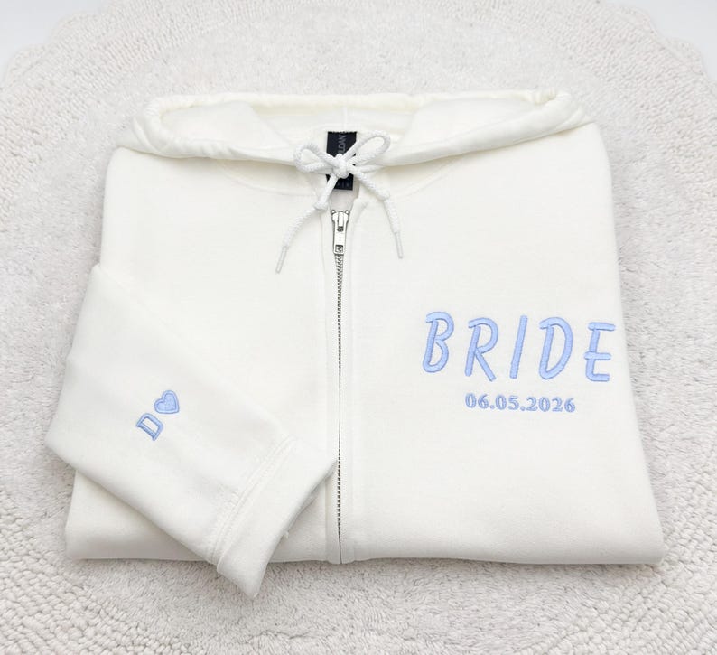 May include: A white zip-up hoodie with the word "BRIDE" embroidered in light blue on the front. The date "06.05.2026" is embroidered below the word. A small blue heart and the letter "D" are embroidered on the sleeve.