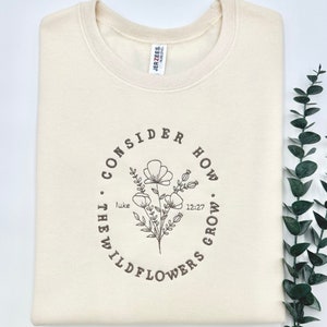 May include: A cream-colored sweatshirt with a brown graphic design that says "Consider how the wildflowers grow" with a floral illustration and the text "Luke 12:27".