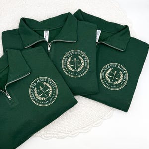 May include: Three dark green quarter-zip pullovers. Each pullover features a white embroidered emblem with the text "CHARLOTTE MOCK TRIAL" and the year "EST. 2013". The pullovers have a ribbed collar and a silver zipper.