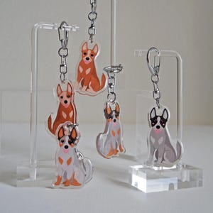 May include: Acrylic dog keychains in various poses and colors, including brown, gray, and white. Each keychain features a silver chain and clasp. The keychains are displayed on clear acrylic stands.