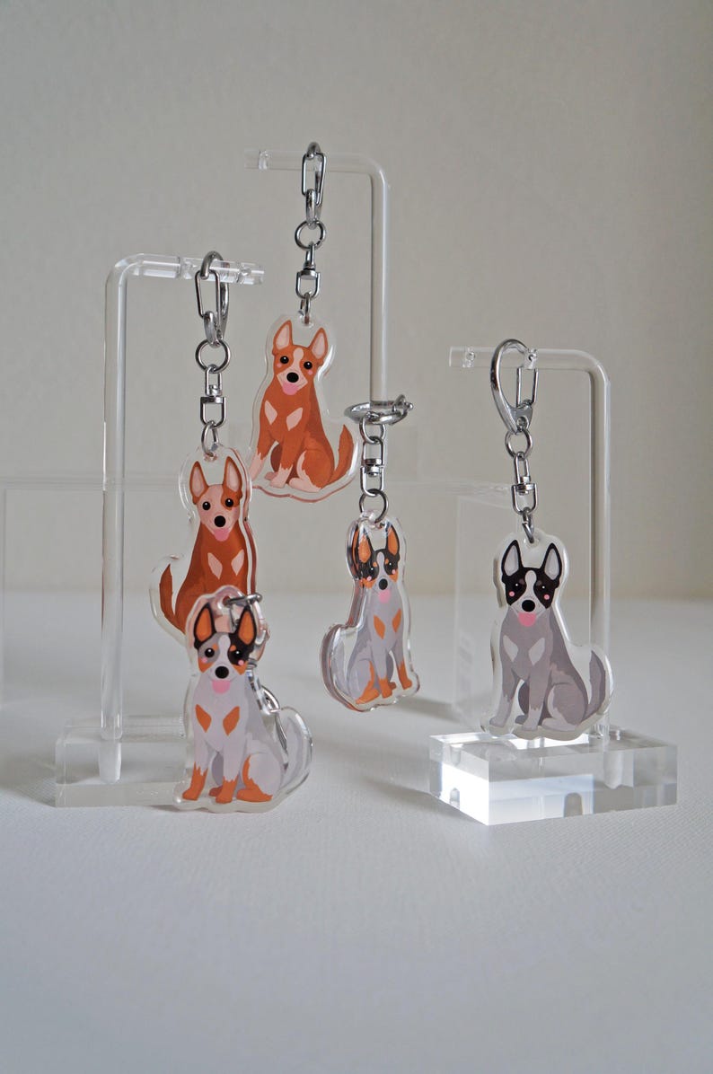 May include: Acrylic dog keychains in various poses and colors, including brown, gray, and white. Each keychain features a silver chain and clasp. The keychains are displayed on clear acrylic stands.