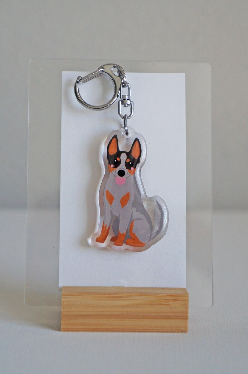May include: A clear acrylic keychain featuring a cartoon illustration of a blue heeler dog. The dog has gray fur, orange markings, and a pink tongue. The keychain has a silver clasp and is displayed on a white card.