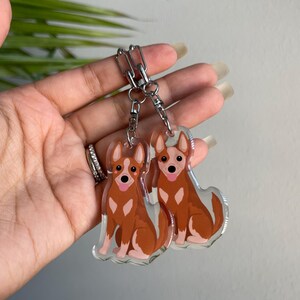 May include: Two clear acrylic keychains featuring a cartoon dog design. The dogs are brown and white with pink tongues, attached to a silver chain. The keychains are held in a hand.