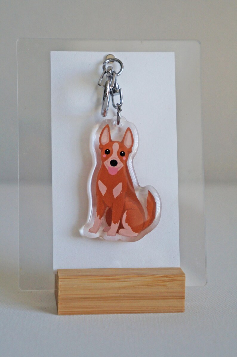 May include: A clear acrylic keychain featuring a cartoon-style dog in shades of brown and white. The dog is sitting with its tongue sticking out. The keychain has a silver-colored metal clasp.