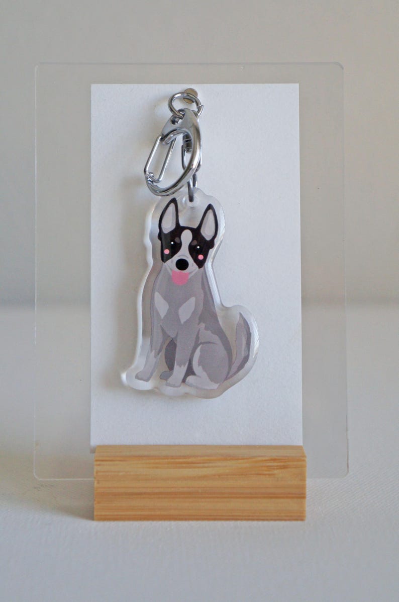 May include: A clear acrylic keychain featuring a seated dog illustration. The dog is gray with black markings around the eyes and ears, a pink tongue, and a silver clasp. The keychain is displayed on a white card and wooden base.