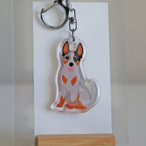 May include: A clear acrylic keychain featuring a cartoon-style Australian Cattle Dog. The dog is depicted in shades of gray, orange, and black, with a silver keyring attached. The keychain is displayed on a white background.