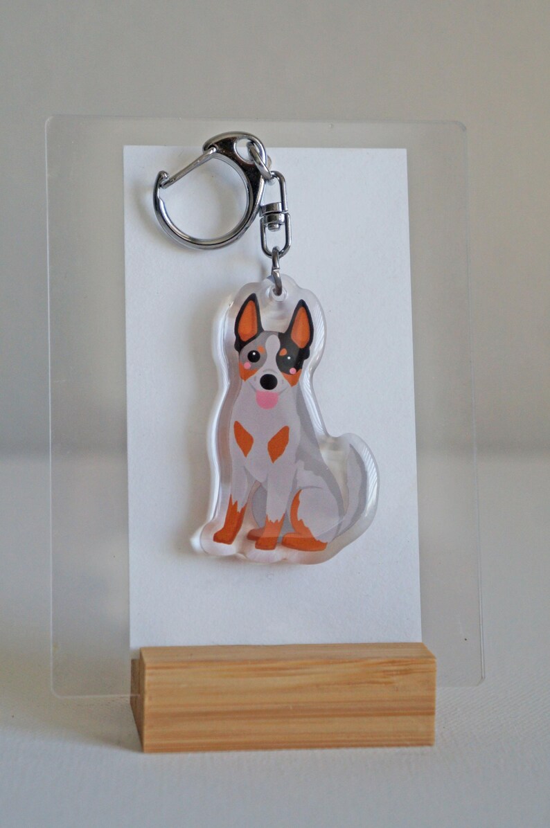 May include: A clear acrylic keychain featuring a cartoon-style Australian Cattle Dog. The dog is depicted in shades of gray, orange, and black, with a silver keyring attached. The keychain is displayed on a white background.