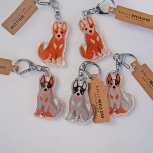 May include: Keychains featuring dog illustrations in orange and gray, with silver key rings and brown tags. The tags read "kiwi & WILLOW KEYCHAINS". The dogs are in a sitting position.