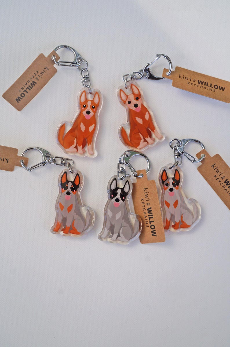 May include: Keychains featuring dog illustrations in orange and gray, with silver key rings and brown tags. The tags read "kiwi & WILLOW KEYCHAINS". The dogs are in a sitting position.