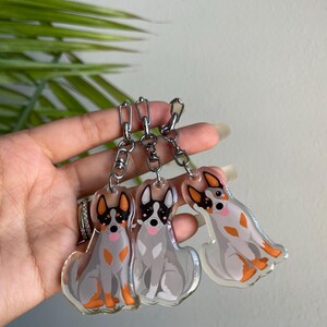 May include: Three clear acrylic dog keychains with silver chains. Each keychain features a cartoon dog design with gray, orange, and black markings. The dogs are sitting with their tongues out. The keychains are attached to a silver chain.