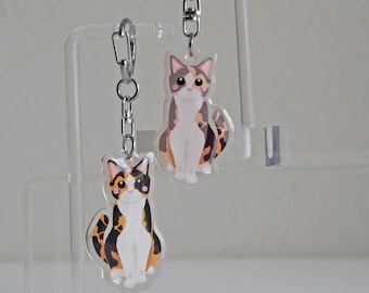 Puede incluir: Dos llaveros de gato de acrílico transparente. Cada llavero presenta un diseño de gato de dibujos animados con un cuerpo blanco y parches de pelaje marrón, negro y naranja. Los llaveros están sujetos a cierres metálicos plateados.