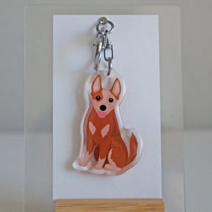 May include: A clear acrylic keychain featuring a cartoon-style dog in shades of brown and white, with a pink tongue. The keychain hangs from a silver chain and is displayed on a white card and a wooden stand.