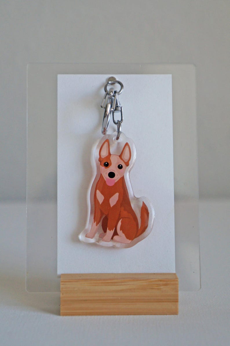 May include: A clear acrylic keychain featuring a cartoon-style dog in shades of brown and white, with a pink tongue. The keychain hangs from a silver chain and is displayed on a white card and a wooden stand.