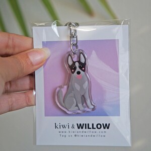 May include: A clear acrylic keychain featuring a cartoon dog with gray fur, black and white markings, and a pink tongue. The keychain is attached to a silver metal clasp. The packaging includes the text "kiwi & WILLOW" and a website address.